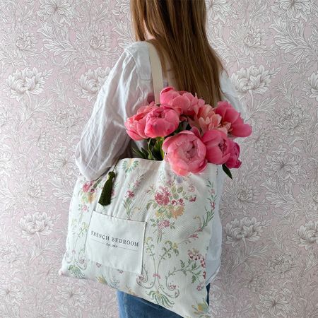 Woman holding a floral tote bag filled with pink peonies, standing against a pink floral wallpaper background.