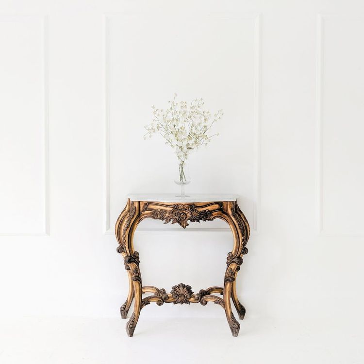 Ornate wooden table with intricate carvings, holding a vase of delicate white flowers, set against a minimalist white paneled wall.