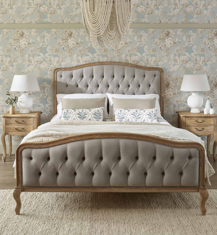 Elegant bedroom with a tufted bed, floral wallpaper, matching wooden nightstands, white lamps, and a chandelier above.