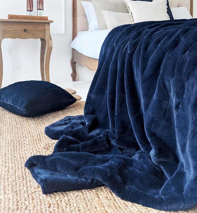 A plush navy blue blanket draped over a bed with a matching pillow on a woven rug, next to a wooden side table.