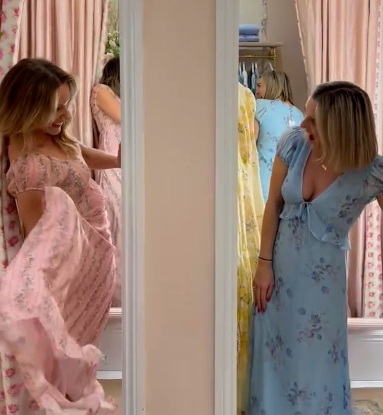 Two women in a fitting room trying on floral dresses, one in pink and the other in blue, smiling and posing in front of mirrors with pink curtains.
