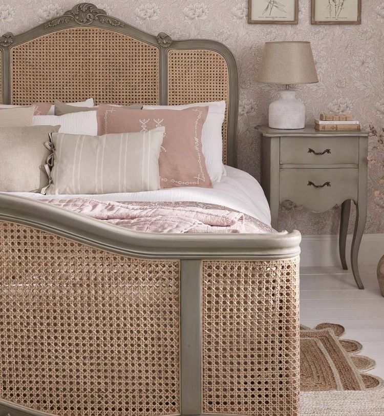 Elegant bedroom with a cane bed, pastel pillows, a vintage nightstand, lamp, and floral wallpaper creating a cozy, classic ambiance.