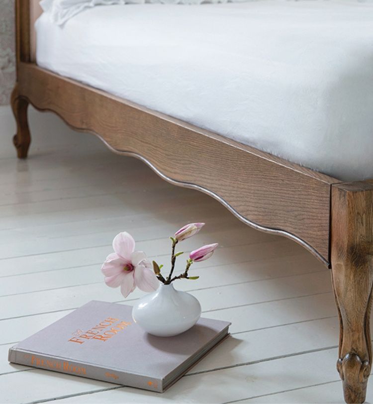 A wooden bed with a white mattress on a light floor. A small vase with pink flowers and a book titled "French Room" are nearby.