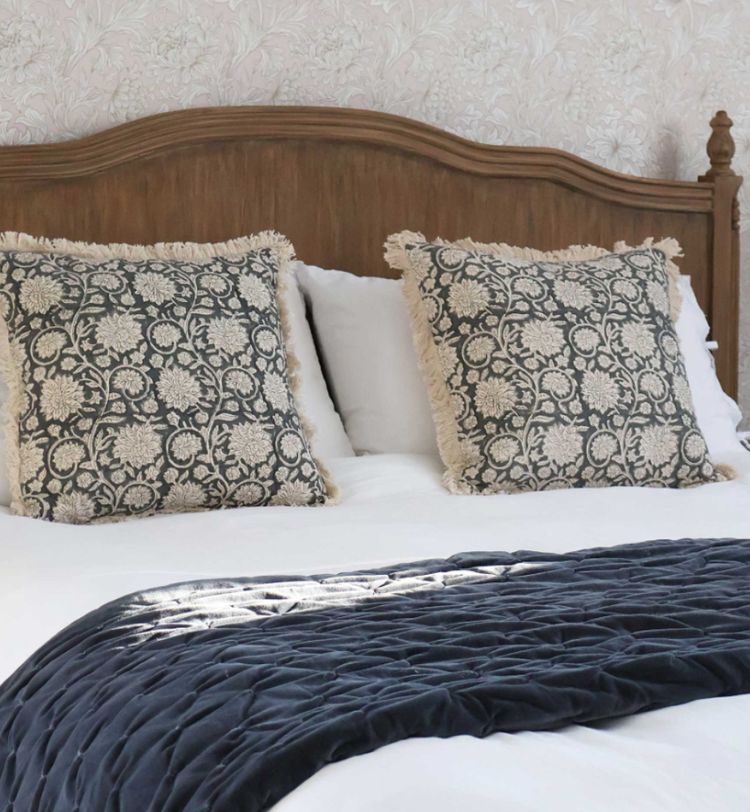 A neatly made bed with a wooden headboard, two patterned cushions, and a dark blue quilted throw in a softly lit room.