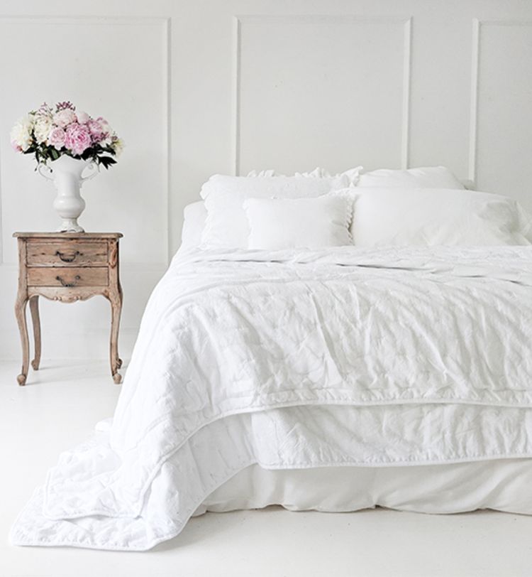 A white bed with fluffy pillows and a quilt, next to a wooden nightstand with a vase of pink and white flowers against a white paneled wall.