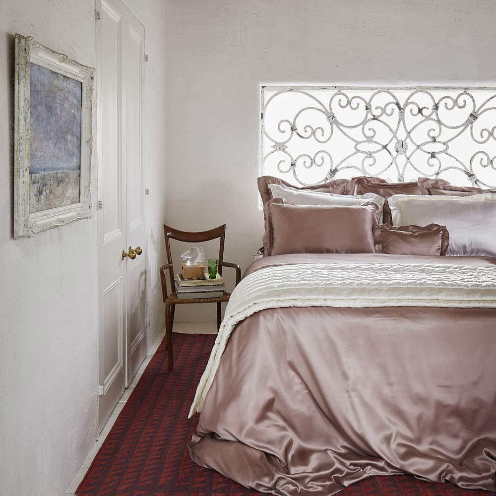 Elegant bedroom with a satin bedspread, decorative iron window panel, wooden chair with books, and a framed painting on a white wall.