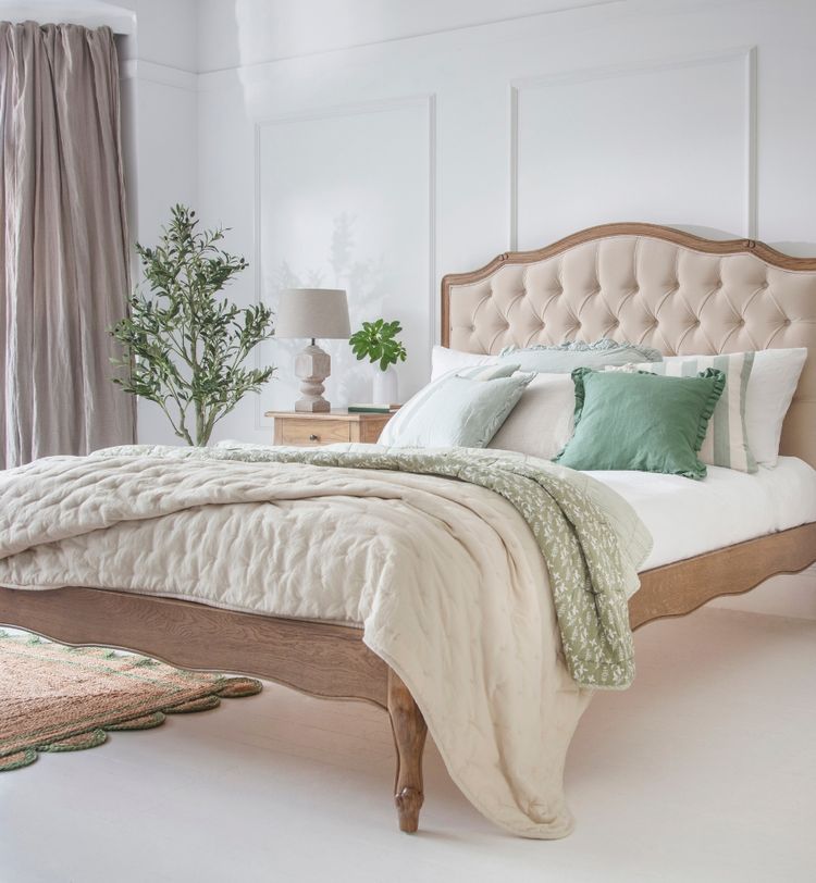 Elegant bedroom with a wooden bed, tufted headboard, green and white pillows, and a cozy quilt. A plant and lamp sit on a nightstand by the window.