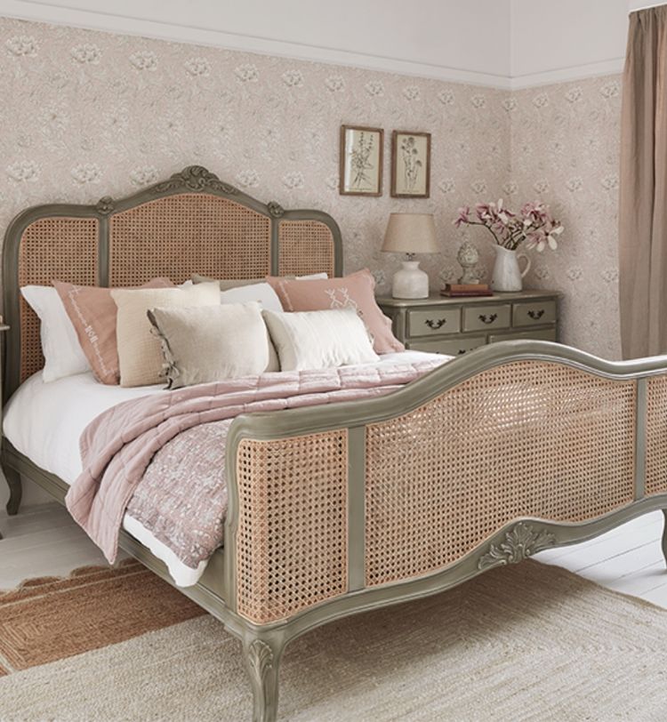 Vintage-style bedroom with a wicker bed, pink and beige bedding, floral wallpaper, wooden dresser, decorative frames, and a vase of flowers.