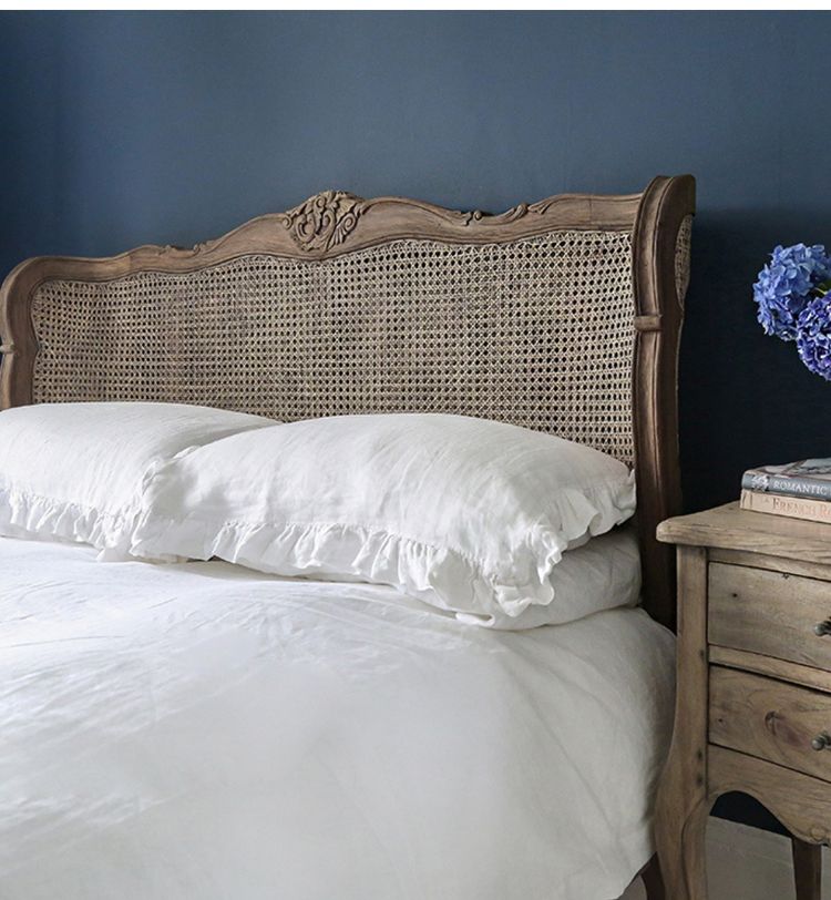 Wooden bed with white linens and a wicker headboard against a dark blue wall. A wooden nightstand holds books and a vase of purple flowers.