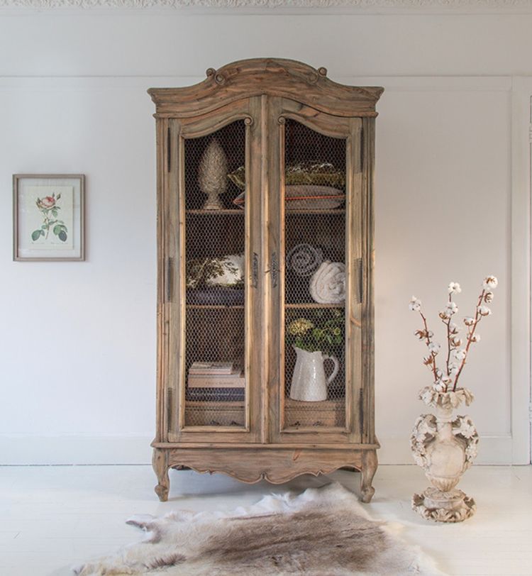 Vintage wooden cabinet with mesh doors, displaying decor items. Framed flower print on wall, ornate vase with branches, and rug on white floor.
