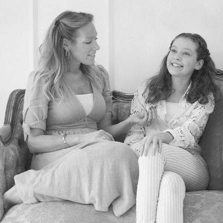 A woman and a young girl sit on a vintage sofa, smiling and talking. The woman wears a light dress; the girl wears a lace top and striped pants.