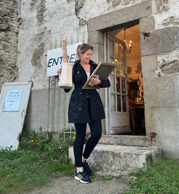 Woman in black coat and sneakers stands at a rustic shop entrance, holding a vase and framed picture, with a chandelier visible inside.