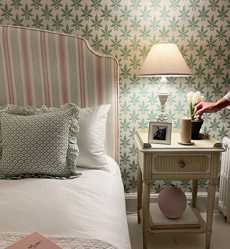 Cozy bedroom with a striped headboard, green patterned pillow, bedside table with a lamp, framed photo, candle, and a hand adjusting a plant.