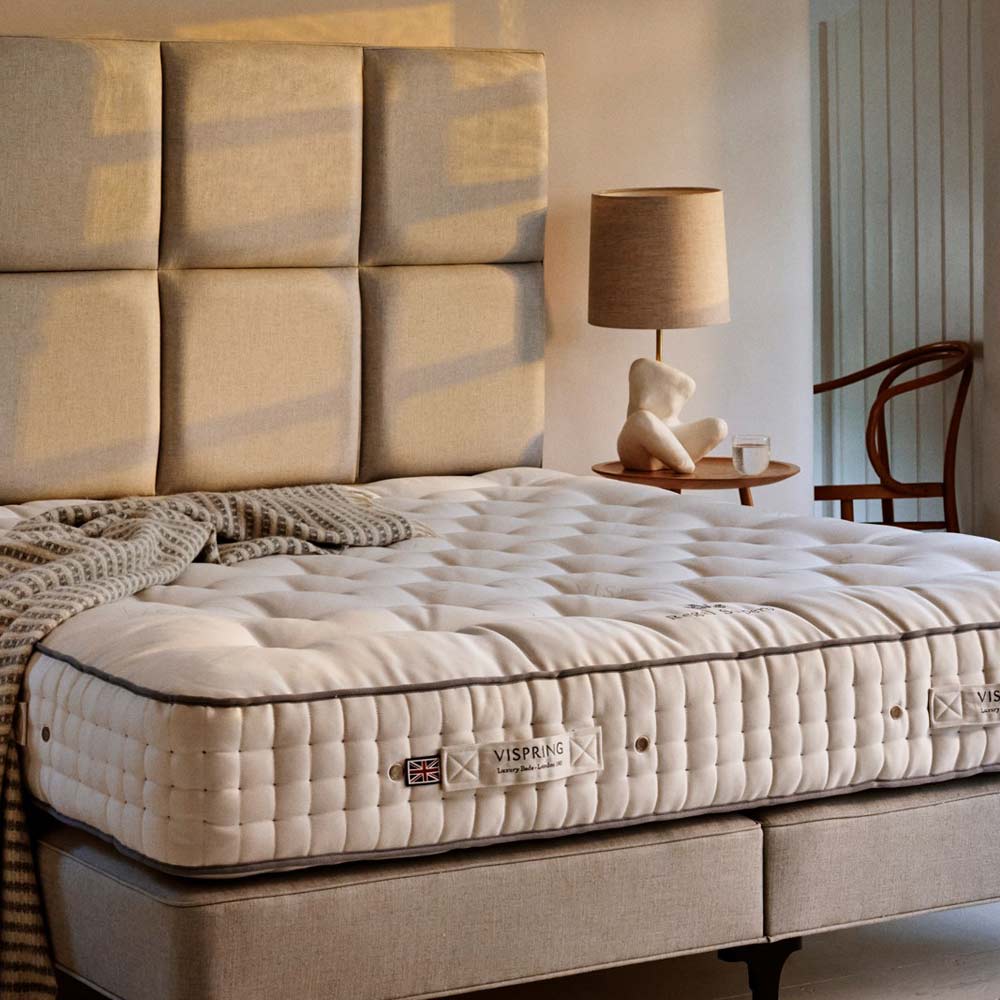 Luxurious bed with a tufted mattress, beige headboard, and a cozy knitted blanket, beside a wooden chair and a lamp on a side table.