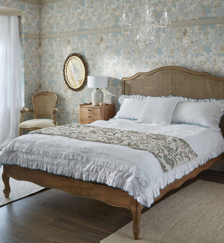 Elegant bedroom with floral wallpaper, ornate chandelier, wooden bed with white bedding, vintage chair, round mirror, and a window with sheer curtains.