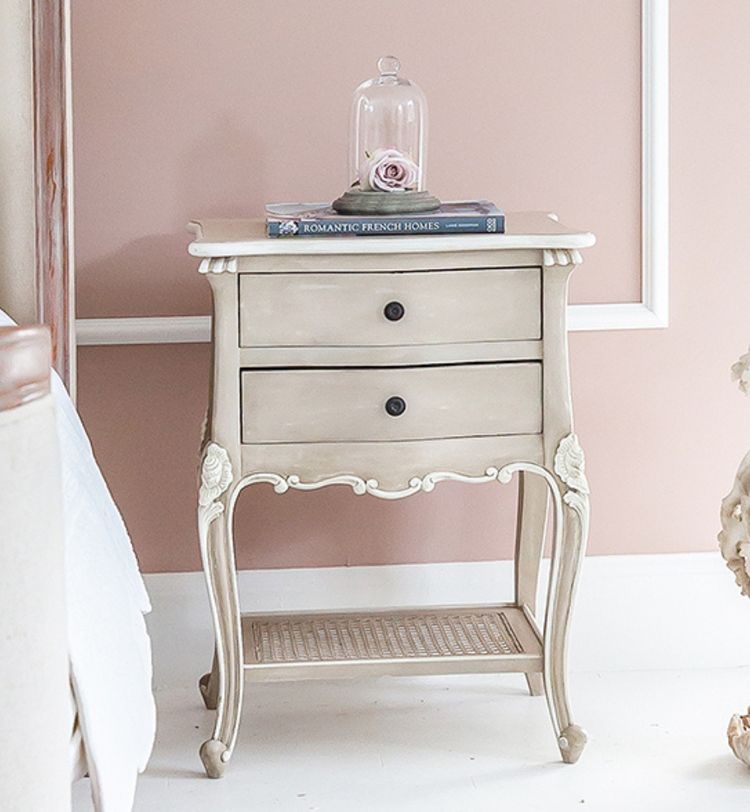 Vintage-style beige nightstand with two drawers, a book, and a glass dome containing a rose on top, set against a pink wall.