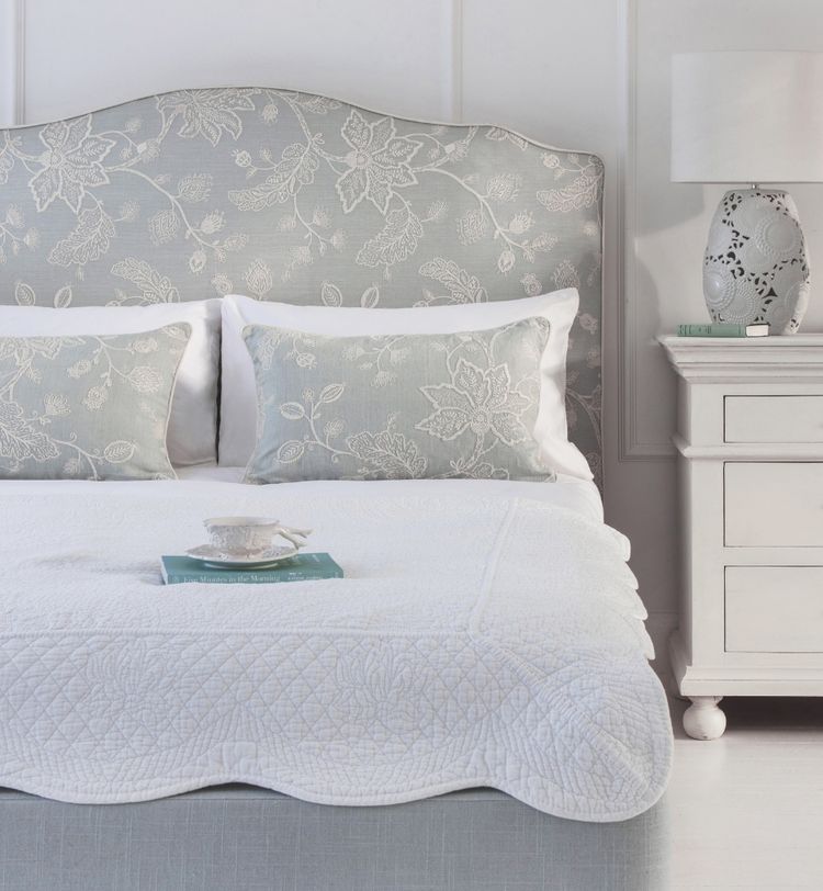 Elegant bedroom with a light blue floral headboard, matching pillows, white quilt, bedside table with lamp, and a teacup on a book.