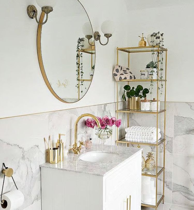 Elegant bathroom with marble accents, gold fixtures, round mirror, and shelves holding plants, towels, and decor items.