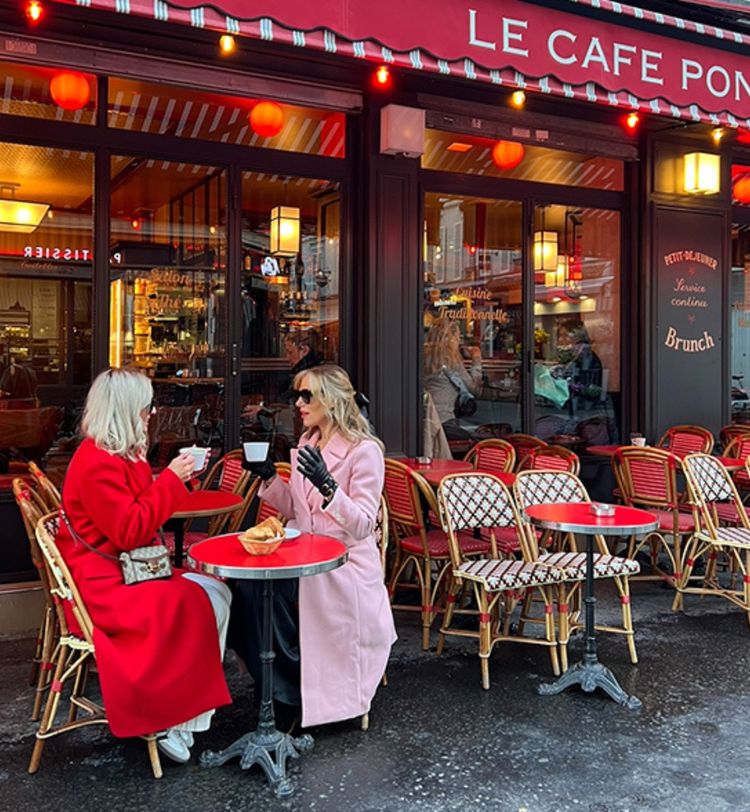 Two women in red and pink coats enjoy coffee at an outdoor café with red awning and wicker chairs.