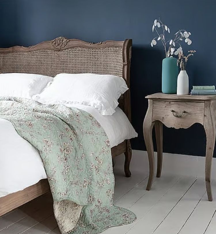 Cozy bedroom with a wooden bed, floral quilt, white pillows, and a vintage nightstand holding vases against a dark blue wall.