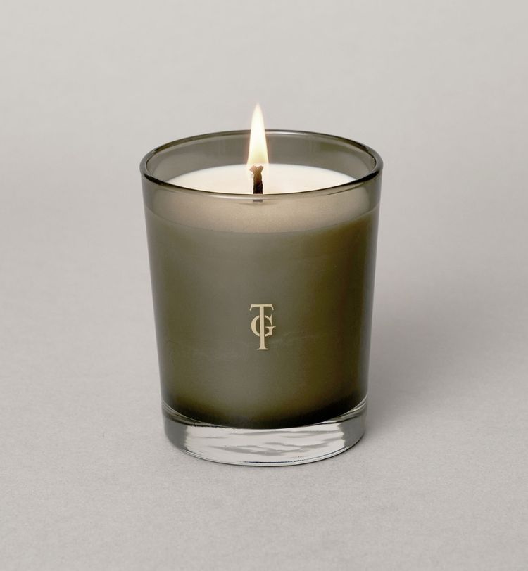 A lit candle in a dark glass holder with a gold monogram on the front, placed on a light gray surface.