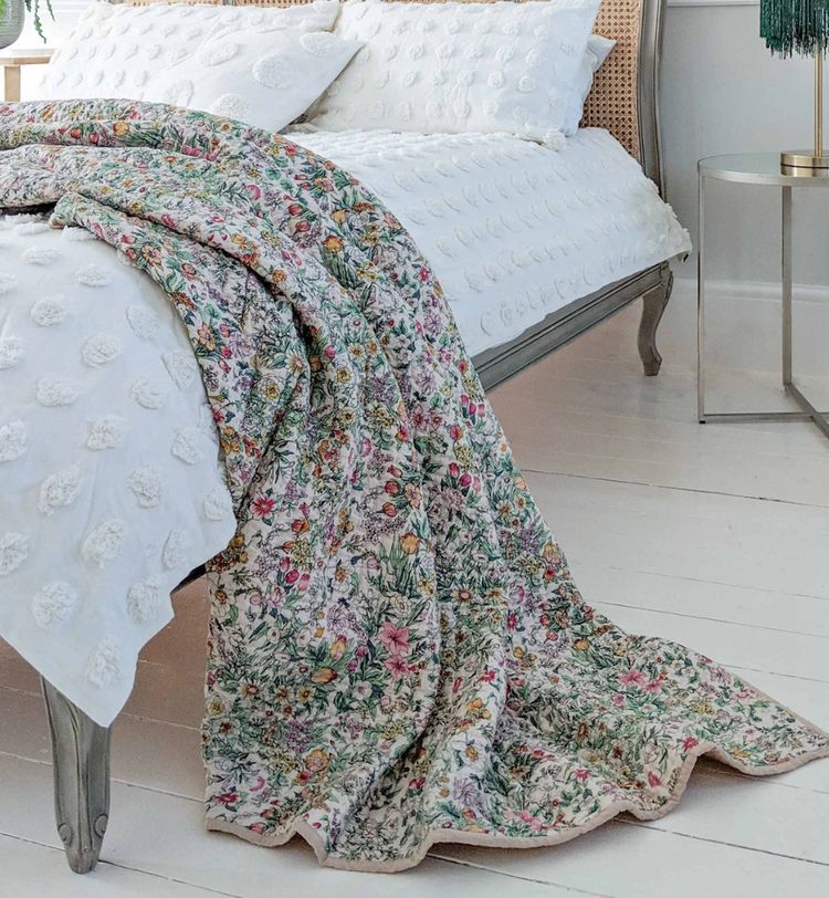 Floral quilt draped over a white bed with textured pillows, next to a round metal side table on a white wooden floor.