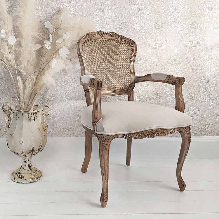 Elegant wooden chair with a woven back and cushioned seat beside a decorative vase with pampas grass, set against a floral wallpaper.