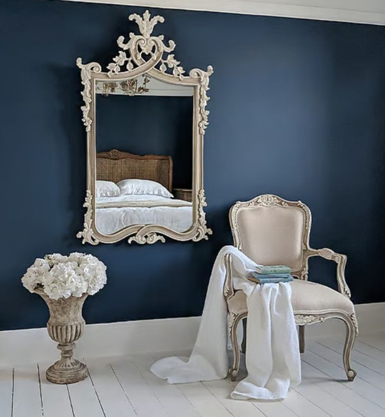 Elegant room with a vintage mirror reflecting a bed, ornate chair draped with a towel, and a vase of white flowers, set against a navy wall.