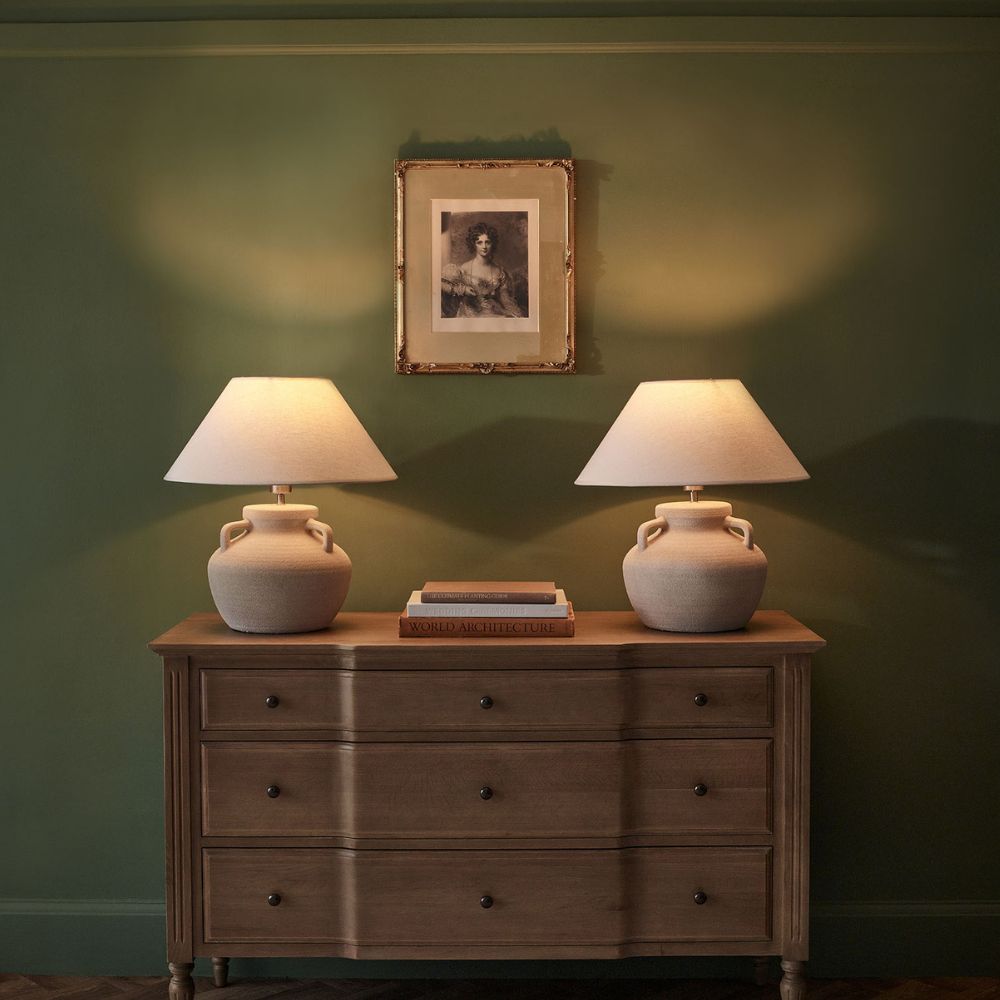 A wooden dresser with two lamps and books is against a green wall. A framed black-and-white portrait hangs above, illuminated by the lamp light.