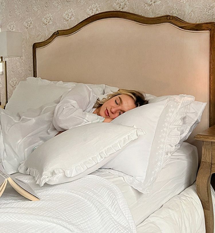 Woman sleeping peacefully in a cozy bed with white bedding and ruffled pillows, a book open beside her on a wooden nightstand.