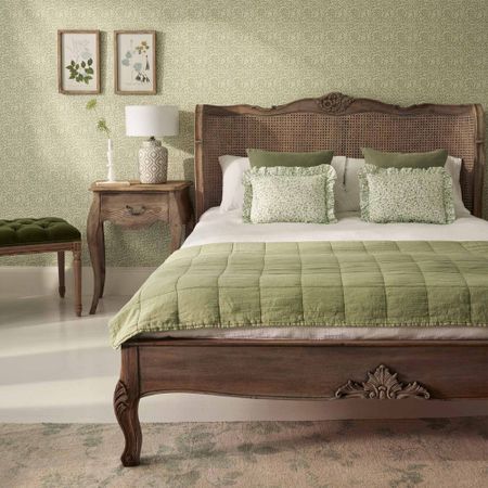 Bedroom with a vintage wooden bed, green quilt, floral pillows, matching wallpaper, nightstand with lamp, and framed botanical prints.