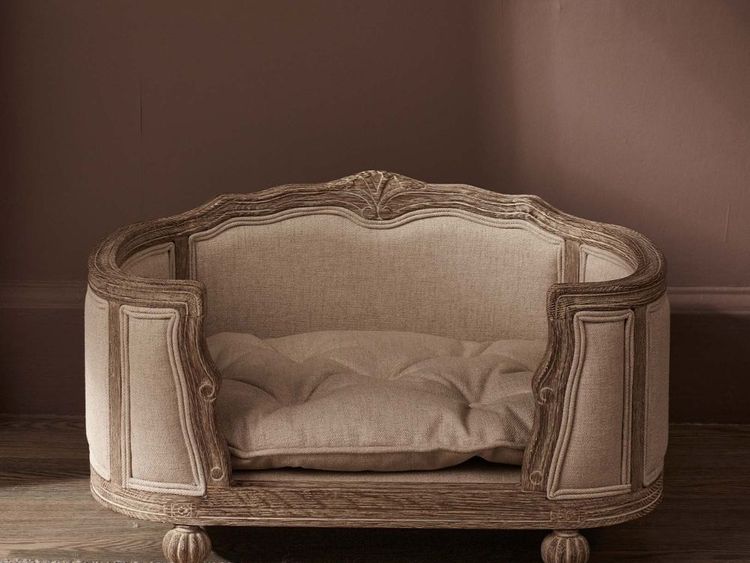 Elegant vintage-style pet bed with ornate wooden frame, tufted beige cushion, and rounded legs, set against a warm-toned wall.