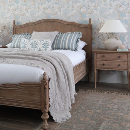 A cozy bedroom with a wooden bed, striped and patterned pillows, a beige throw, a wooden nightstand, and floral wallpaper.