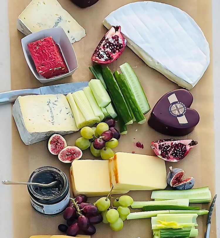 A cheese board with various cheeses, figs, grapes, cucumber sticks, pomegranate, and a jar of jam, arranged on brown paper.