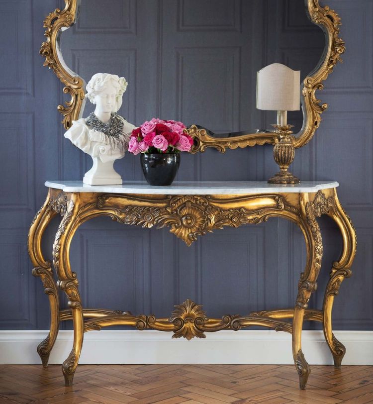 Elegant gold console table with ornate mirror, topped with a white bust, pink roses in a black vase, and a small lamp.