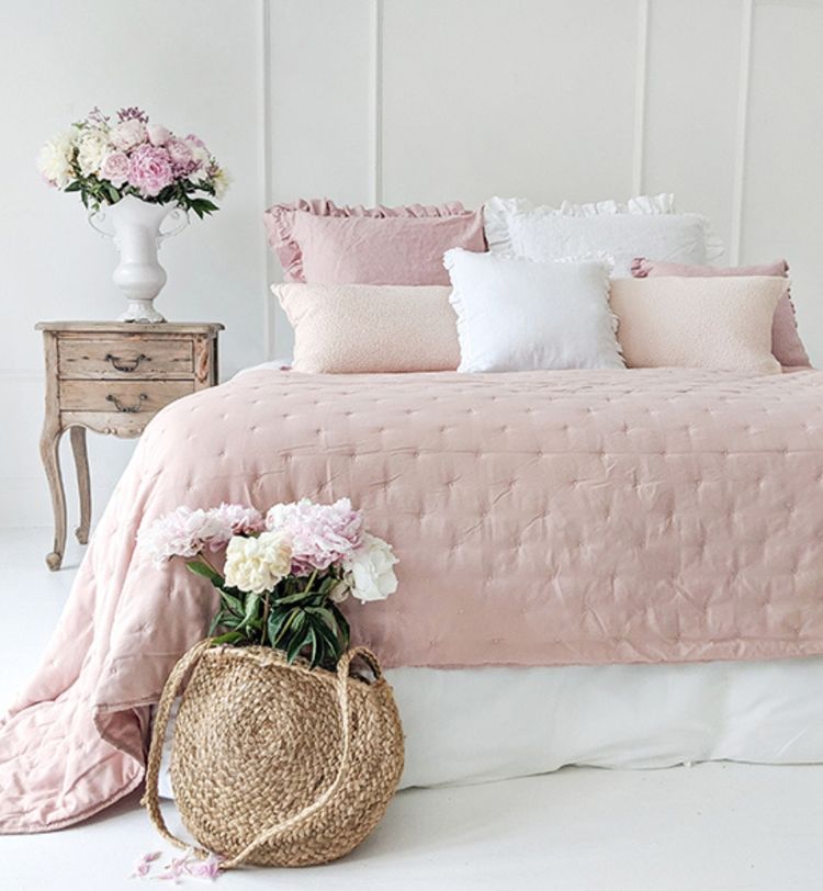 A cozy bedroom with a pink quilted bedspread, white and pink pillows, a rustic nightstand, and vases of pink and white flowers.