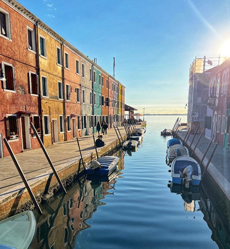Colorful buildings line a canal in bright sunlight, with small boats moored along the waterway and a clear blue sky above.