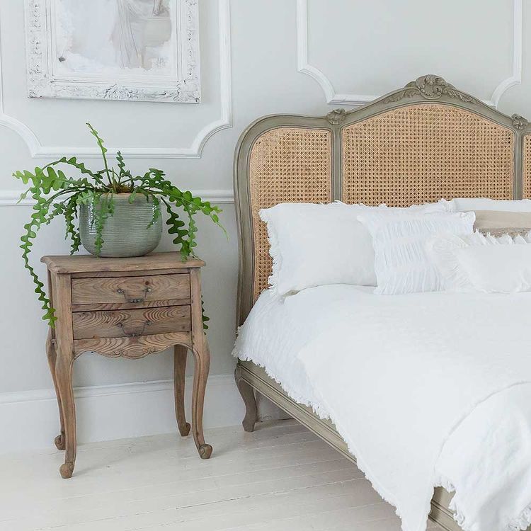 Elegant bedroom with a vintage wooden bed, white bedding, and a rustic nightstand holding a potted fern. Light gray walls and a framed artwork.
