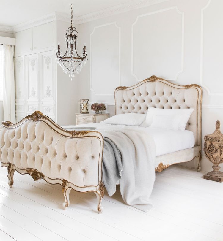 Elegant bedroom with a tufted bed, ornate chandelier, white walls, and wooden floors. Neutral tones create a luxurious, serene atmosphere.