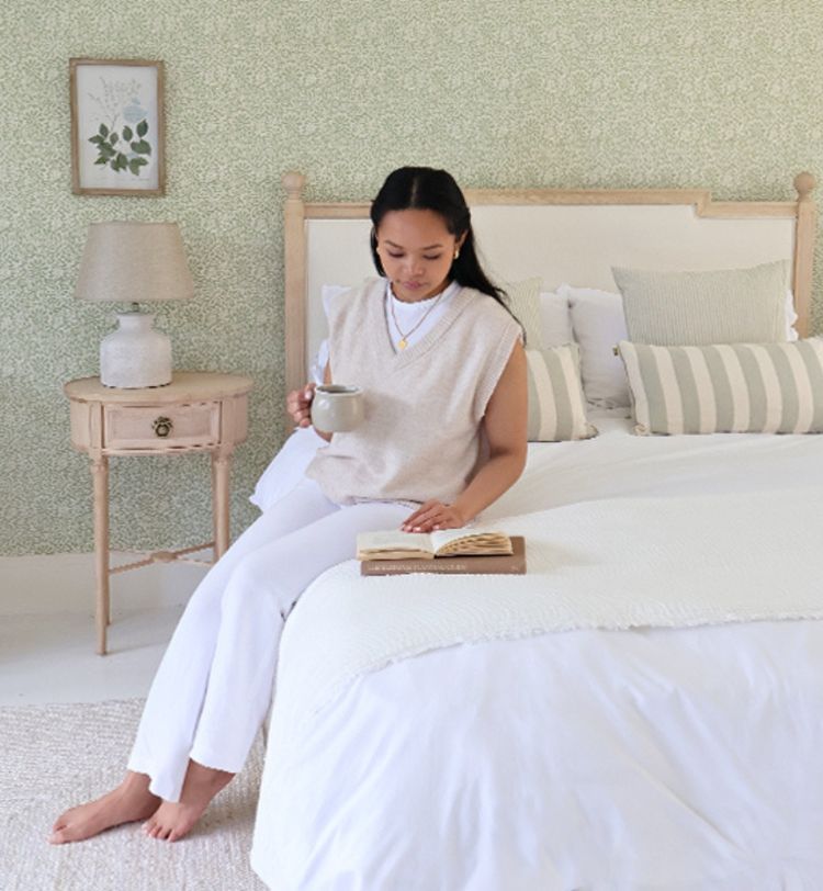 A woman sits on a bed, holding a mug and reading a book. The room has a green patterned wall, a bedside table, and a lamp.