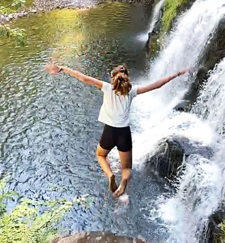 Person jumping into a pool from a waterfall, arms outstretched, surrounded by lush greenery and cascading water.