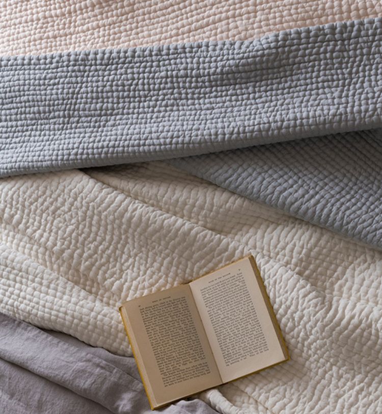 Open book on a bed with textured, layered blankets in shades of pink, gray, and cream. Cozy and inviting atmosphere.