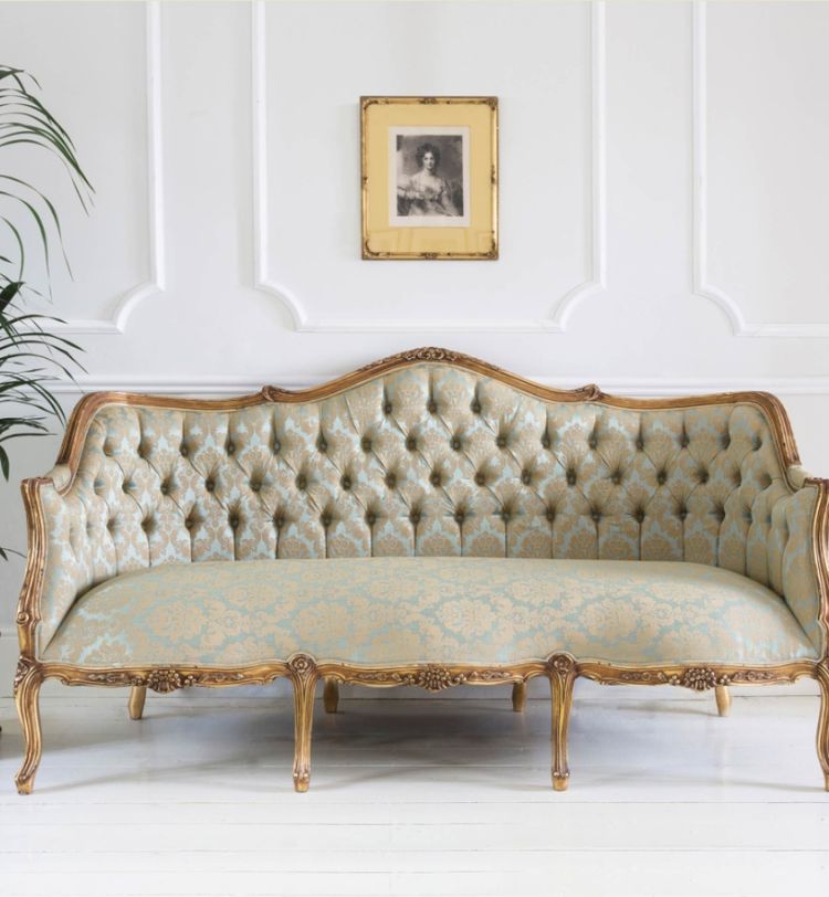Elegant vintage sofa with ornate wooden frame and tufted upholstery in a classic room. Framed portrait and potted plant in the background.
