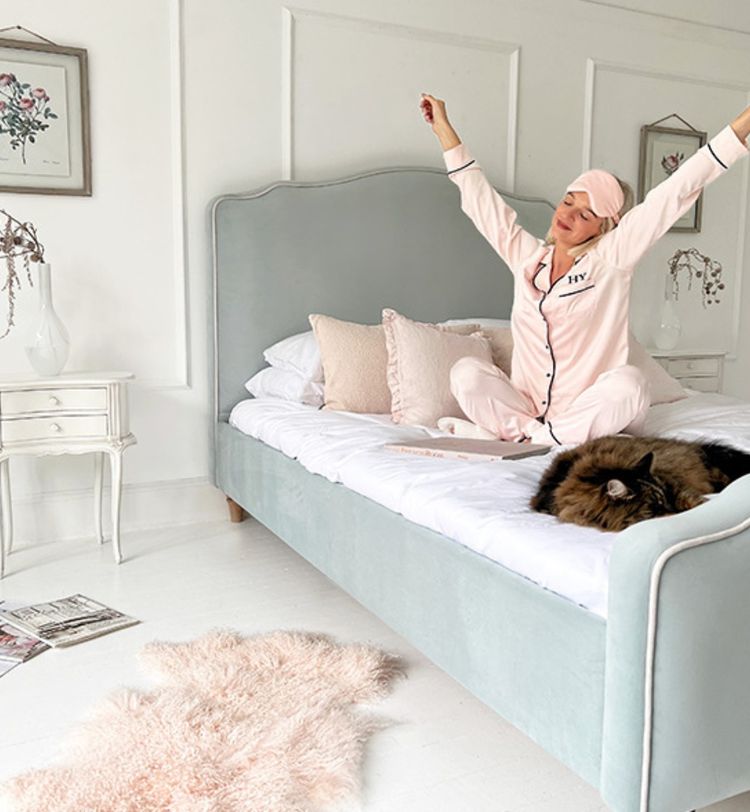Woman in pink pajamas stretches on a bed with a fluffy cat. The room is white with elegant decor and a soft rug on the floor.