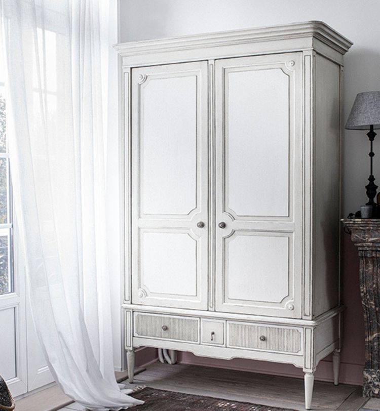 White vintage wardrobe with paneled doors and two drawers, beside a window with sheer curtains and a dark lamp on a mantel.