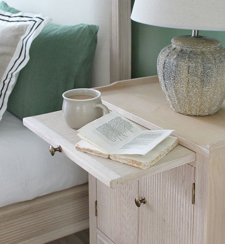 A cozy bedroom scene with a wooden bedside table, a mug, an open book, and a textured lamp beside a bed with green and white pillows.