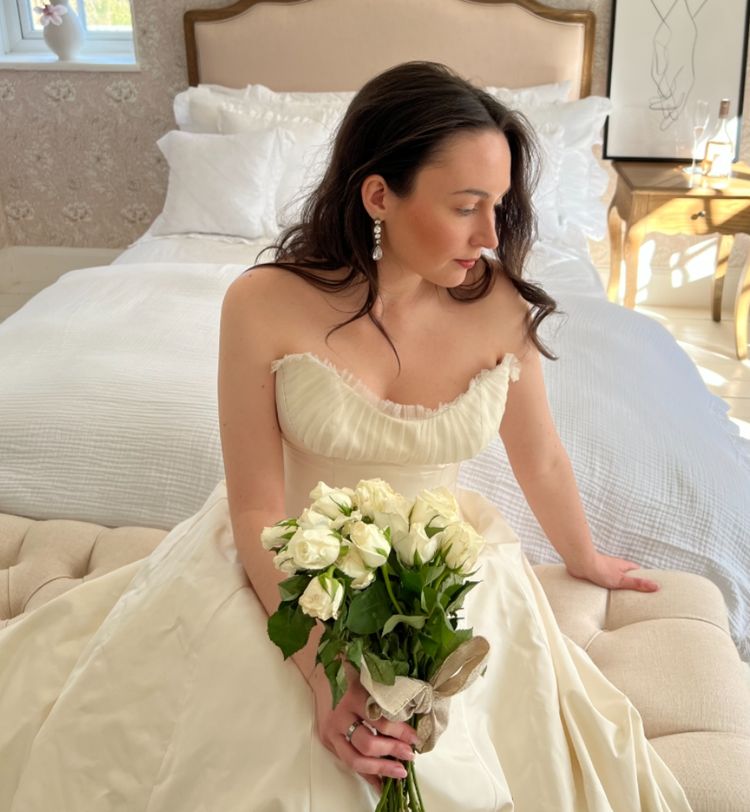 Bride in a strapless gown sits on an ottoman holding white roses, with a bed and soft lighting in the background.