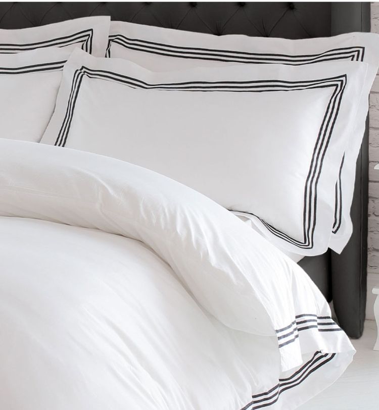 White bedding set with black striped borders, featuring two pillows and a duvet on a tufted dark headboard, next to a white nightstand.