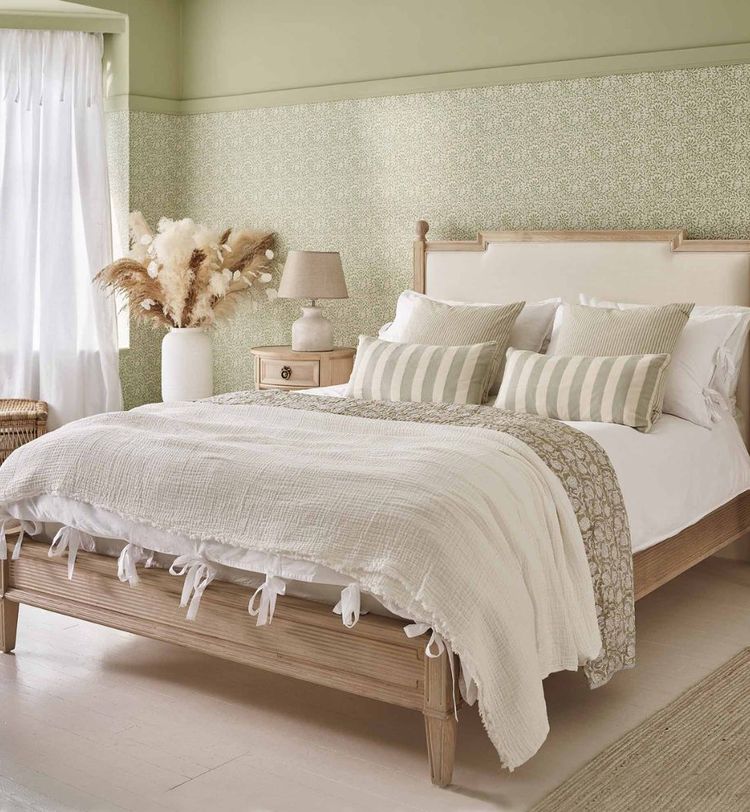 Cozy bedroom with a wooden bed, white and beige bedding, striped pillows, a nightstand with a lamp, and a vase of dried flowers by the window.