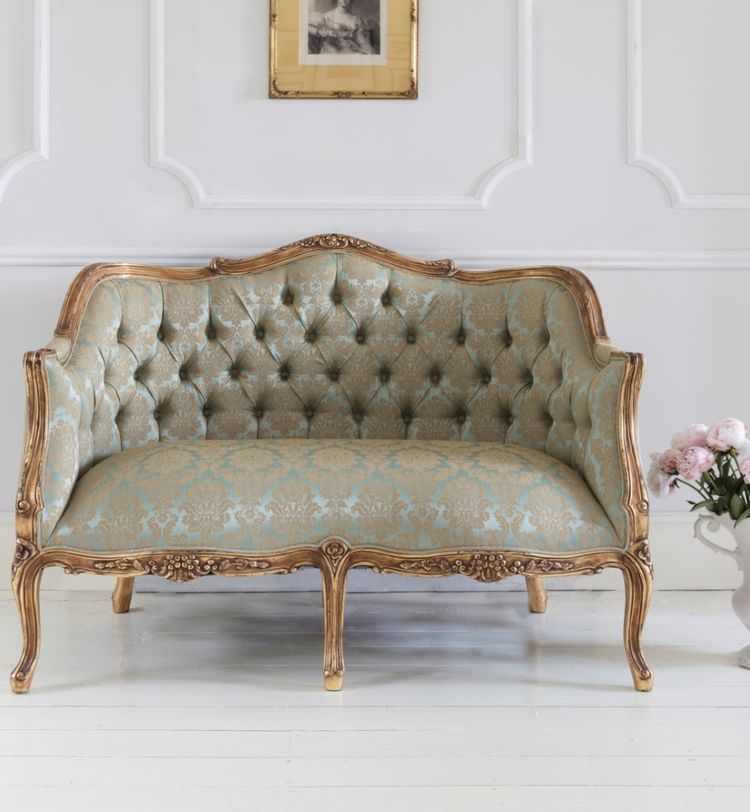 Elegant vintage loveseat with ornate wooden frame and tufted floral upholstery, set against a white paneled wall with a framed picture above.
